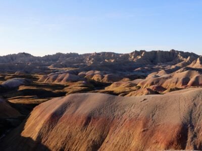 late afternoon sun casts long shadows across undulating badlands national park ridges in muted earth tones of beige, rust, ochre, and gray, with sparse dry grass in valleys under a clear blue sky.