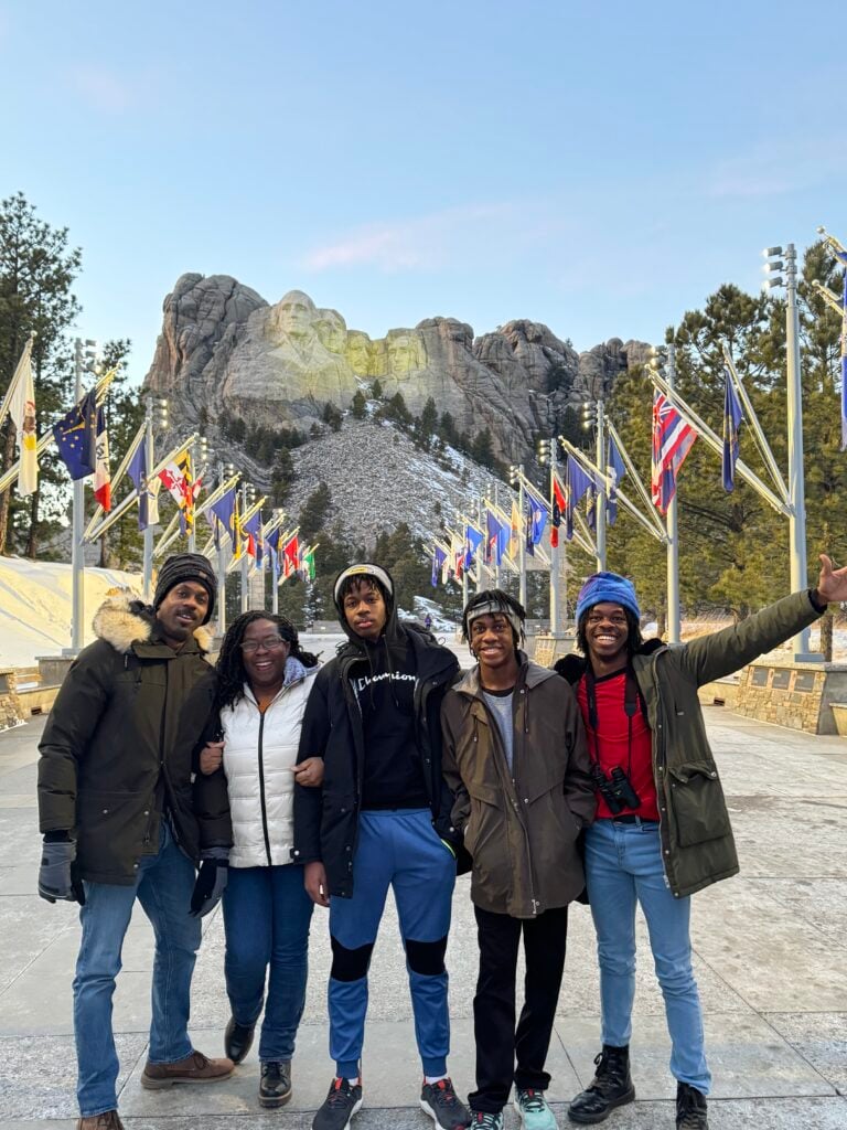 Family of five in winter jackets standing arm-in-arm on a stone promenade with colorful flags and snow-dusted Mount Rushmore in the background.