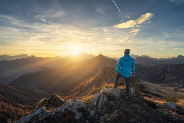 A lone hiker in a bright blue jacket stands on a rocky mountain crest at sunrise, overlooking layered peaks and wispy clouds in an alpine-like landscape.
