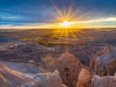 sunrise over the badlands in south dakota, with golden rays illuminating layered rock formations against a lavender sky.