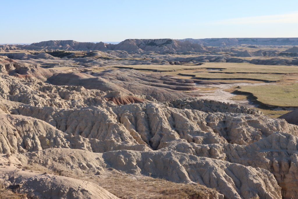 South Dakota Sightseeing: A Personal Journey 6 panoramic view of undulating eroded clay and sandstone badlands formations in soft beige, cream, and muted rose hues beneath a clear pale blue sky.